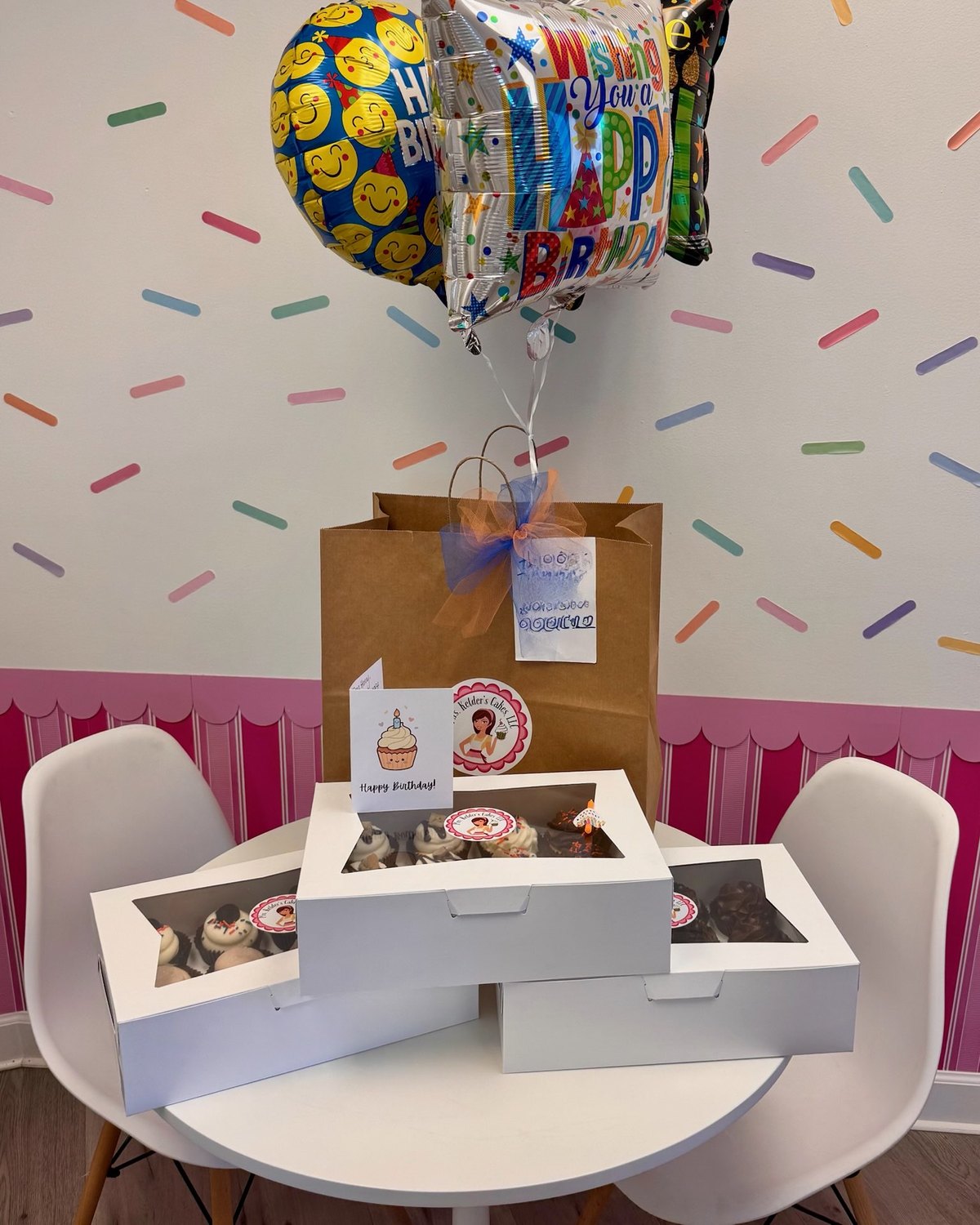 A campus delivery ready to go out: a kraft gift bag with a tulle bow and handwritten tag, birthday balloons and boxes of Mrs. Kelder's cupcakes