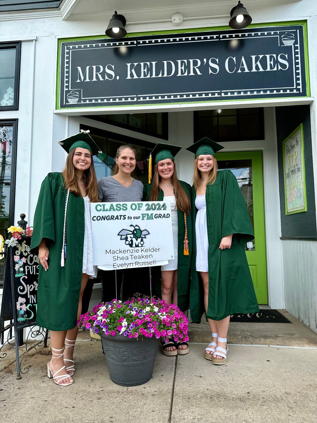 Class of 2024 graduates in green gowns outside the Mrs. Kelder's Cakes sign